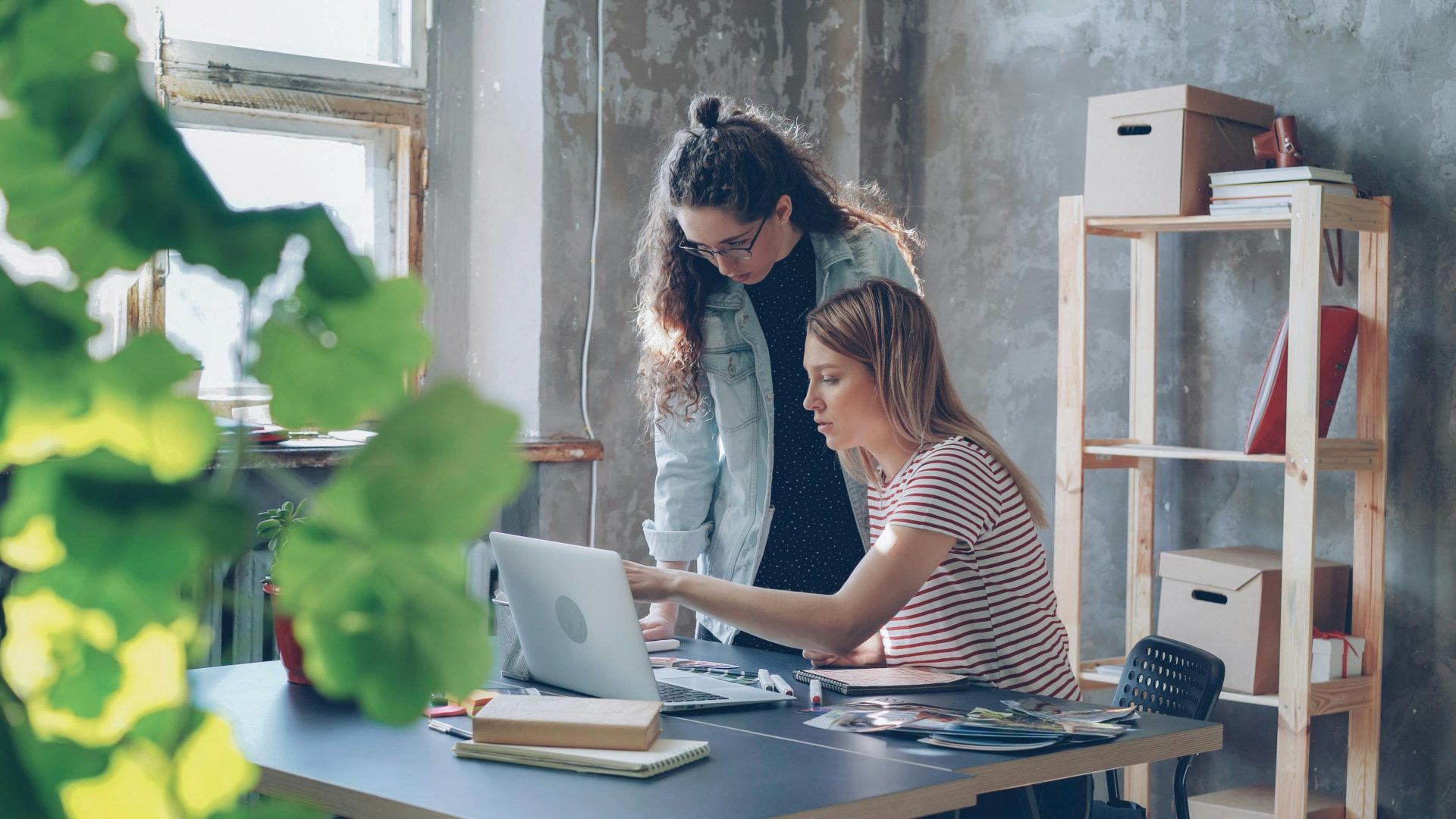 Zwei junge Frauen arbeiten gemeinsam in einem modernen Büro mit Laptop und Pflanzen.