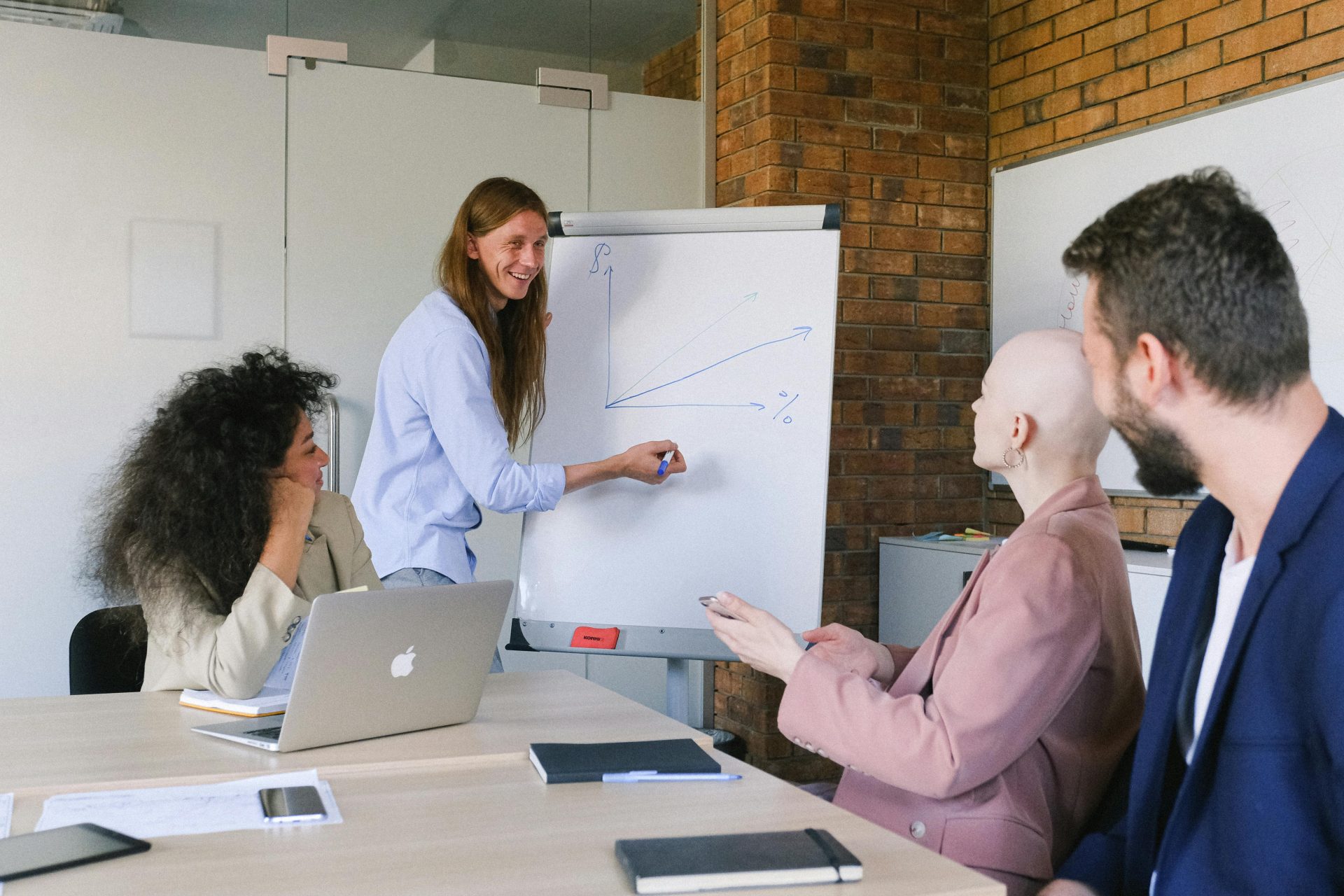 Ein lächelnder Sprecher zeichnet tagsüber in einem modernen Arbeitsumfeld ein Diagramm auf ein Whiteboard und erklärt seinen Kollegen dabei die Details eines Projekts.